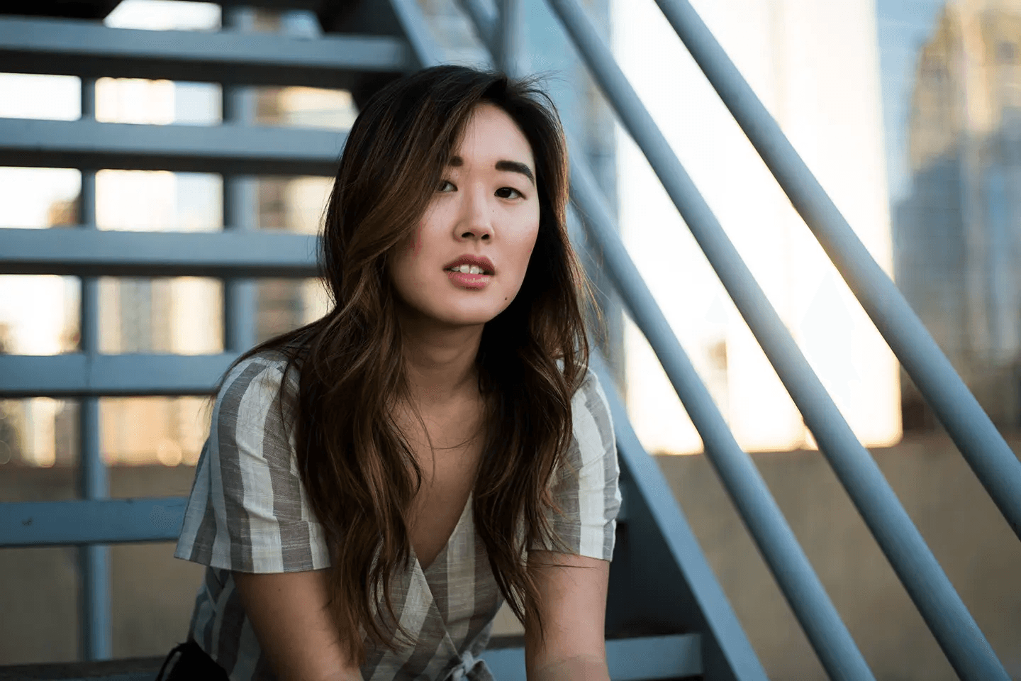 A young woman with long, wavy hair gazes directly at the camera while sitting on a metal staircase. The soft lighting and neutral tones create a calm and contemplative mood. The striped pattern of her shirt adds a touch of visual interest to the portrait.